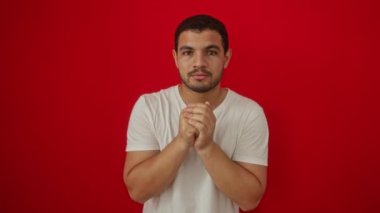 Beyaz tişört giyen genç İspanyol adam başarı ve zafer için heyecanlı kollarını kaldırmış, izole edilmiş kırmızı arka planda gururlu.
