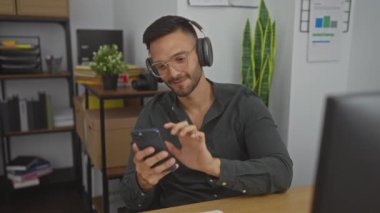 Sakallı, kulaklık takan ve gülümseyen genç adam modern ofiste arka planda bitki ve rafları olan telefonları kontrol ederken.