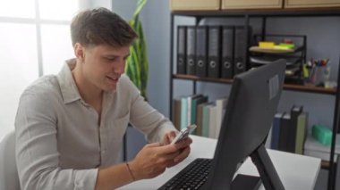 Ofiste gülümseyen genç bir adam arka planda ofis malzemeleri ve masada bilgisayarını kullanırken akıllı telefonunu kullanıyor.