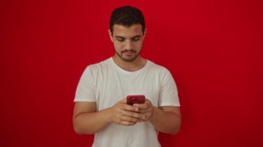 Akıllı telefon kullanırken mutlu gülümseyen genç İspanyol adam izole edilmiş kırmızı arka planda olumlu onay işareti veriyor.