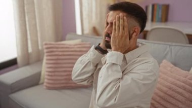 Rahat bir oturma odasında oturan genç adam, elleri yüzü üzerinde, yumuşak yastıklar ve kitaplıklarla çevrili, tefekkür ya da stresi tasvir eden.