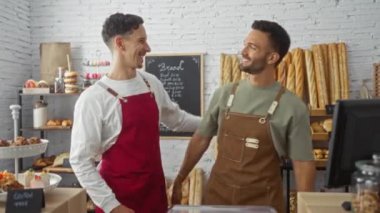 Biri kırmızı önlüklü, diğeri kahverengi önlüklü iki yetişkin adam, arka planda çeşitli ekmek ve hamur işleri olan bir fırında gülümserken sarılıyorlar..