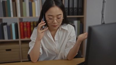 Ofis ortamında telefonla konuşan kadının endişeli bir ifadesi var. Arkaplanda kitap rafları ve dosyalar var. Bu da genç, Çinli bir profesyonel için stresli bir iş durumu anlamına geliyor.