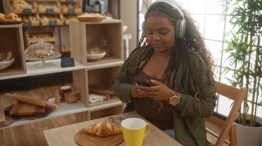 Afro-Amerikalı bir kadın fırında akıllı bir telefonla kahvenin tadını çıkarıyor. Etrafı fırın malzemeleriyle çevrili..