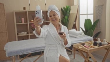 Spa merkezinde orta yaşlı bir kadın bir bardak şarabın tadını çıkarırken akıllı telefonuyla selfie çekiyor..