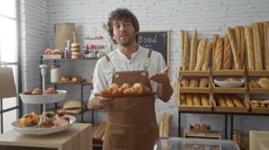 Fırındaki genç adam çeşitli ekmek ve hamur işleriyle dolu rafların önünde dururken taze kruvasanlardan bahsediyor.