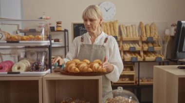 Pastanede çalışan bir kadın. Etrafı ekmek rafları ve çöreklerle çevrili.