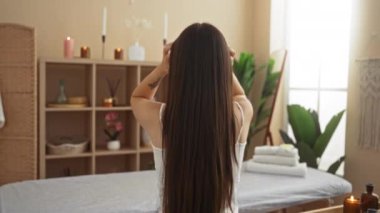 Uzun saçları olan bir kadın, mumlar ve bitkilerle çevrili, sakin bir spa odasında ellerini tarayıp evin içinde huzur verici bir sağlık atmosferi yaratıyor.