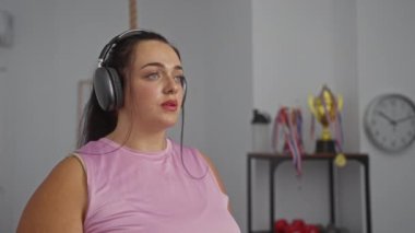 Spor salonunda egzersiz yapan, kulaklık takan ve arka planda kupaları ve saati olan bir kadın.