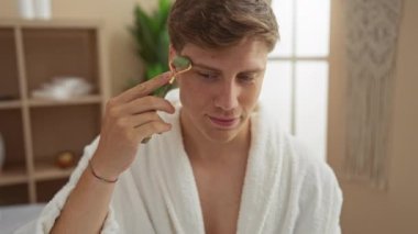 Genç adam spa odasında yüz masajı için yeşim taşı kullanıyor bornoz giyerken, kapalı bir güzellik merkezinde sağlık ayinleri sergiliyor.