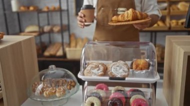Fırında elinde kahve ve bir tabak kruvasan olan yakışıklı genç adam, önlük takıyor, hamur işleriyle dolu bir tezgahın arkasında duruyor, içeride çeşitli fırınlanmış mallar sergiliyor.