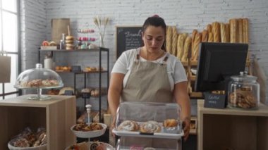 Fırında kıvrımlı bir kadın tezgahın arkasındaki hamur işlerine bakıyor önlük takıyor, etrafı fırınlanmış ekmeklerle, kurabiyelerle, raflarla çevrili.