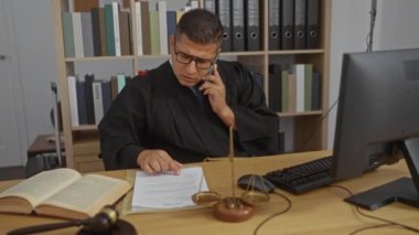 Yargıç cüppeli genç adam telefonla konuşuyor ve hukuk kitapları ve bilgisayarla dolu bir ofiste masasında belgeler okuyor..