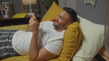 Sakallı genç bir İspanyol adam telefonunu kullanırken gülümsüyor. Yumuşak ışıklandırmalı ve modern bir iç mekanı olan rahat bir yatak odasında sarı bir yastığın üzerinde yatıyor..