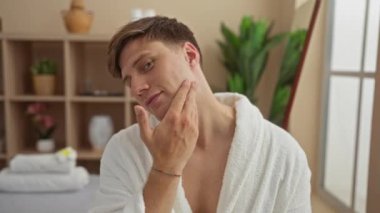 Kaplıca odasında beyaz bir bornoz giymiş yakışıklı bir adam arka planda bitki ve raflarla modern, kapalı bir güzellik salonuna dokunuyor.