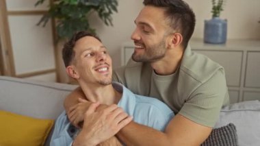 Gülümseyen eşcinsel çift, kanepede oturmuş sıcak oturma odasında birbirlerine sevgi ve beraberlik gösteriyorlar.