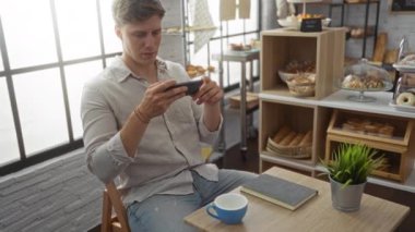 Fırında akıllı telefon kullanan genç adam, arka planda çeşitli fırınlanmış mallarla dolu raflar ve ahşap bir masada defter ve kahve fincanı ile masada otururken...