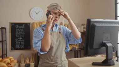 Fırında telefonla konuşan bir kadın bilgisayara bakarken arka planda ekmek ve duvarda bir saatle endişeleniyor.