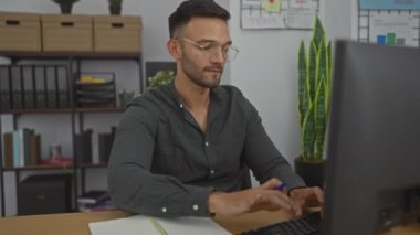 Ofis masasında çalışan genç İspanyol bir adam modern bir iş yerinde bilgisayarla çalışıyor, gözlük takıyor ve deftere yazmaya odaklanıyor..