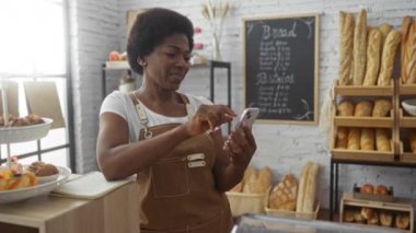 Fırında çalışan bir kadın, tebessümle telefona bakıyor. Etrafı taze ekmek ve hamur işleriyle çevrili. Raflı, modern bir dükkanda.