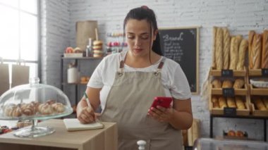 Fırında genç bir kadın elinde telefon, elinde ekmek ve raflarla notlar tutuyor.