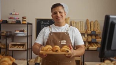 Önlüklü genç İspanyol adam gülümsüyor ve ekmek ve tatlılarla dolu sıcak bir pastanede taze hamur işleri sunuyor..