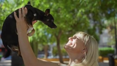 Yeşil ağaçlar ve arka planda bulanık binalar olan şehir parkında küçük siyah bir chihuahua köpeği tutan bir kadın..