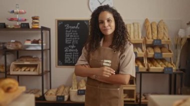 Fırında çalışan kıvırcık saçlı genç bir kadın. Kapalı bir yerde önlük giyiyor ve arka planda raflarda ekmek var.