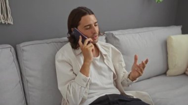 Modern oturma odasındaki bir kanepede otururken akıllı telefondan konuşan İspanyol genç adam.