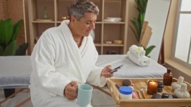 Spa 'da bornoz giymiş genç bir adam elinde fincanla ahşap bir tepside sağIıklı eşyalarla çevrili bir şekilde bir salon ortamında rahatlama ve kendine bakmanın önemini vurguluyor.