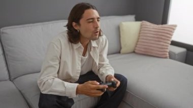 Oturma odasındaki kanepede video oyunları oynayan uzun saçlı genç bir adam.