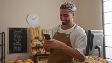 Fırında çalışan genç bir adam, duvarında beyaz bir saat ve arka planda bir ekmek menüsü olan ve etrafında taze ekmek ve hamur işleriyle çevrili bir önlükle telefonunu kullanıyor.