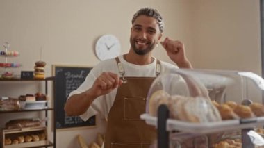 Sakallı genç adam fırında neşeyle dans ediyor. Arkasında ekmek rafları, pastalar ve duvarında bir saat var.