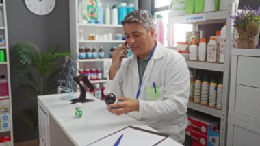 Beyaz önlüklü bir eczacı, iyi stoklanmış bir eczanede telefonla konuşurken ilaç şişesini inceliyor ve tezgahın üzerinde bir pano var..