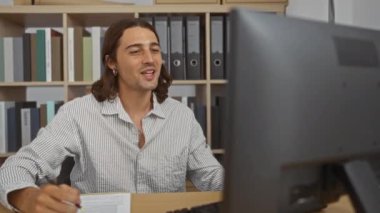 Uzun saçlı yakışıklı genç bir adam bilgisayar ekranına bakarken çalışma odasında notlar alıyor.