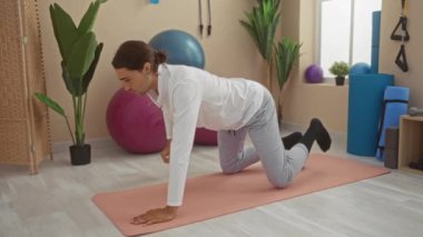 Uzun saçlı genç adam modern bir spor salonunda yoga pozu veriyor. Bitkiler, egzersiz topları, spor aletleri, esneklik ve konsantrasyon göstergesi..