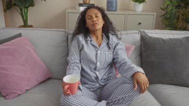 Oturma odasındaki kanepede uzanmış, çizgili pijama giymiş ve elinde pembe bir kupa tutan bir kadın.