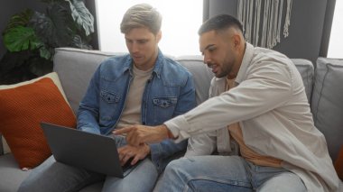 Oturma odasında bitkiler ve yastıklarla sohbet ederken bir adam başka bir adamla koltukta otururken dizüstü bilgisayarı işaret ediyor..