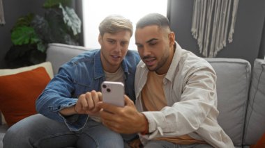 İki yakışıklı adam bir apartman dairesinde oturuyor, birlikte akıllı telefona bakıyorlar, arkadaşlığı ve rahat bir ev ortamını işaret ediyorlar..