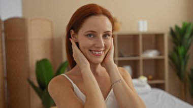 Young redhead woman smiling and touching her face while relaxing in a modern spa salon with indoor wellness setup and plants clipart