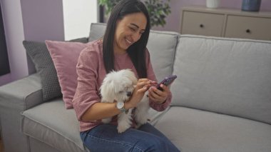 Kapalı bir koltukta oturan gülümseyen bir kadın beyaz bir bizon köpeğini tutuyor ve akıllı telefon kullanıyor..