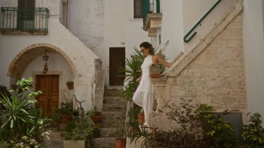 Beyaz elbiseli güzel bir İspanyol kadın Puglia, İtalya 'nın büyüleyici, tarihi sokaklarını keşfediyor. Beyaz taş binalar ve yemyeşil bitkilerle çevrili..