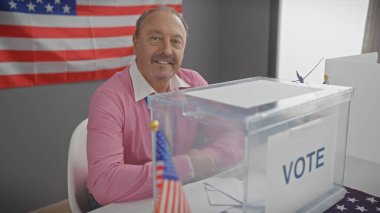 Gülümseyen orta yaşlı bir adam Amerikan bayrağıyla bir odada oturuyor, oy sandığının yanında, ABD 'nin seçmen katılımını resmediyor..