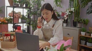 Önlüklü Çinli bir çiçekçi, çiçek buketleri ve bitkilerle çevrili bir çiçekçinin içinde dizüstü bilgisayar ve telefon kullanıyor..