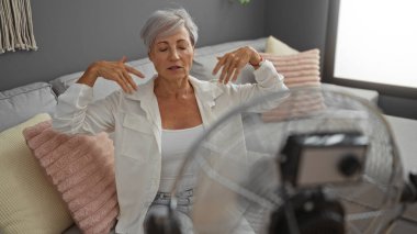 Gri saçlı yaşlı kadın oturma odasında oturuyor ve önünde bir vantilatörle serinliyor, rahat bir koltukta yastıklarla çevrili..