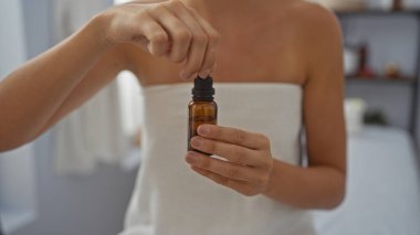 Young woman at spa center holding essential oil bottle indoors in wellness setting with a serene atmosphere clipart