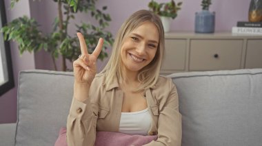 Güzel sarışın bir kadın, bir apartmanın rahat oturma odasında kanepede otururken barış işareti gösteriyor..