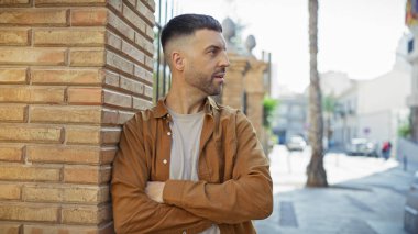 Sakallı yakışıklı İspanyol bir adam güneşli bir şehir sokağındaki tuğladan duvara yaslanıyor..