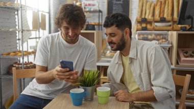 Erkek arkadaşlar birlikte bir fırın kafede oturuyor, bir telefona bakıyor ve içeceklerin tadını çıkarıyor, arkaplanda raflarda hamur işleri sergileniyor.