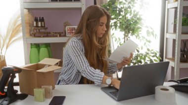 Bir ev dekorasyonu mağazasında dizüstü bilgisayarda çalışan genç bir kadın, paketler ve ürünlerle çevrili, bir masada belgeleri dikkatle inceliyor..